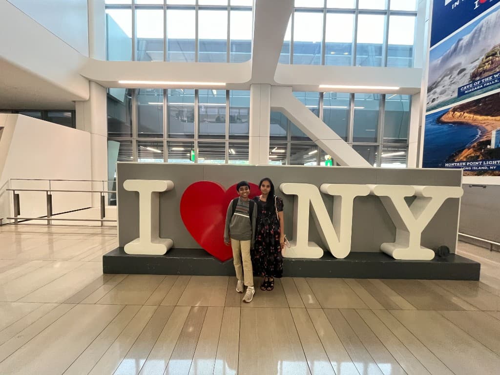 A young visitor and a family member standing in front of a large I Love NY sign in a bright terminal hall.